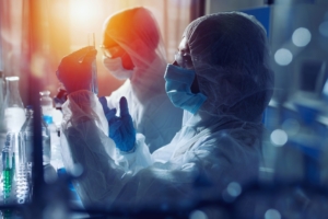 Two people in laboratory suits, face masks, and blue gloves handle a test tube beside lab glassware and liquids.