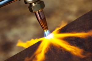 A copper gas cutter nozzle directing blue and orange flames onto a dark metal sheet with a blurred background.