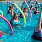 Senior women participate in a water aerobics class, raising colorful pool noodles overhead while exercising in a pool.