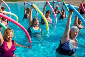 Senior women participate in a water aerobics class, raising colorful pool noodles overhead while exercising in a pool.