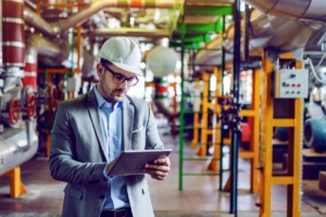 An engineer wearing a hard hat and using a tablet while standing among pipes and machinery in an industrial plant.