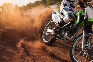 A dirt bike rider turning sharply on a dusty off-road trail as the rear tire kicks up a large cloud of dirt.