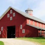 A red barn with open doors, a gravel path, wooden fencing, green grass, and trees under a pale blue sky.