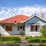 A weathered single-story house has peeling paint, a red metal roof, boarded windows, and an overgrown front yard.