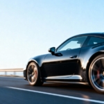 A rear and side view of a black, two-door sports coupe driving on a two-lane highway with a guardrail during the day.