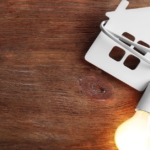 A small piece of wood shaped like a house sitting on a table with a lit light bulb's cord wrapped around it.