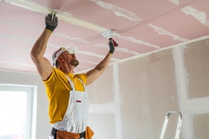 A construction worker in yellow-and-white workwear applies joint compound to drywall seams on a ceiling.