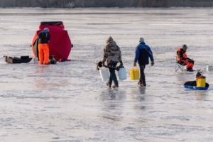 Four people are out on a large, frozen body of water. They are holding fishing poles to go ice fishing.