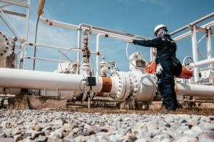 An engineer turning a large, round valve on a long, white pipeline at an oil and gas facility on a sunny day.